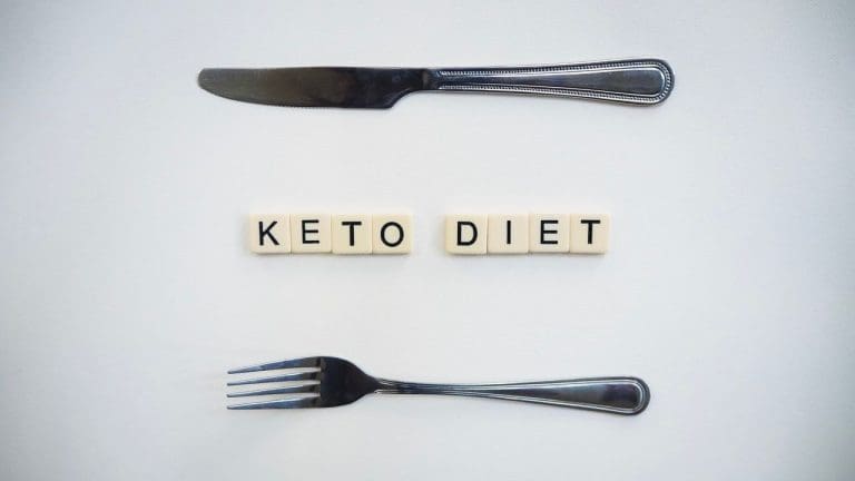 Top view of a knife, fork, and scrabble tiles spelling 'Keto Diet' on a white surface.