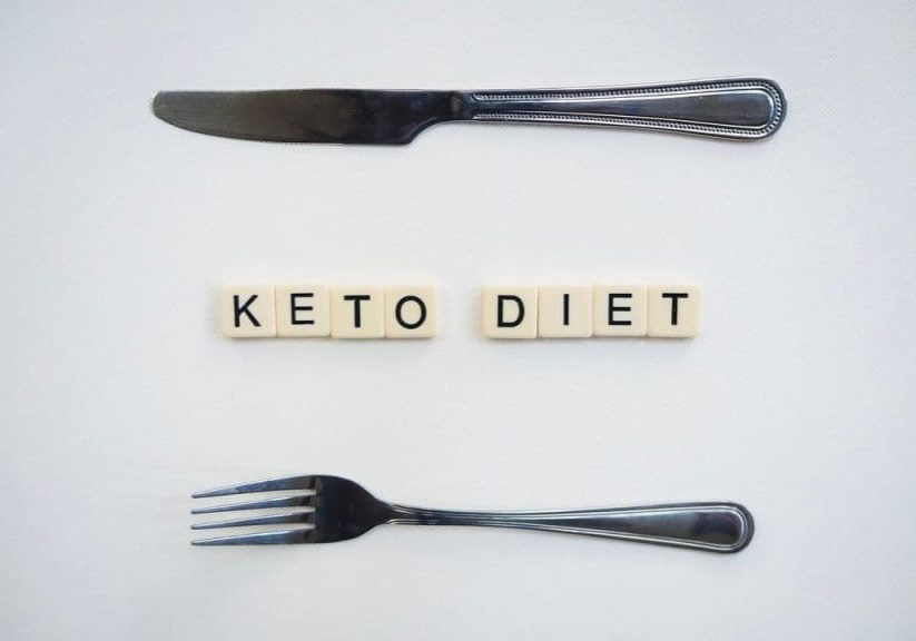 Top view of a knife, fork, and scrabble tiles spelling 'Keto Diet' on a white surface.