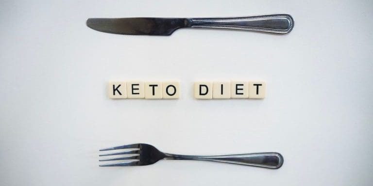 Top view of a knife, fork, and scrabble tiles spelling 'Keto Diet' on a white surface.